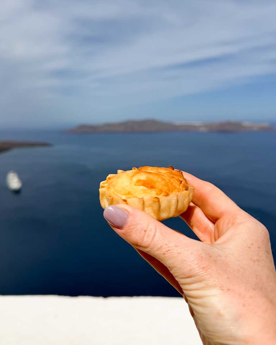 Hand holding a traditional Melitinia pastry overlooking Santorini