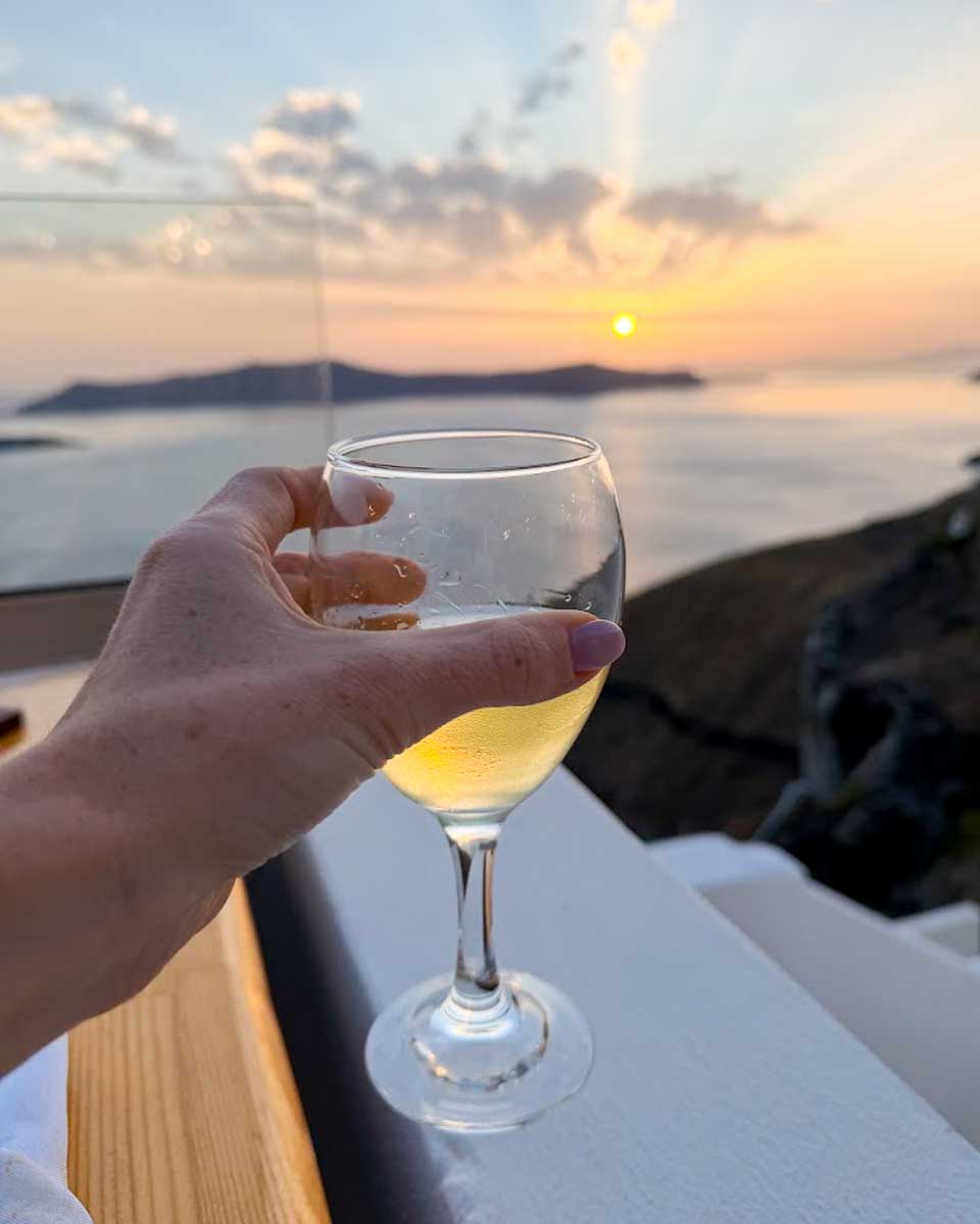 Hand holding a glass of white wine overlooking the Santorini caldera at sunset in Greece