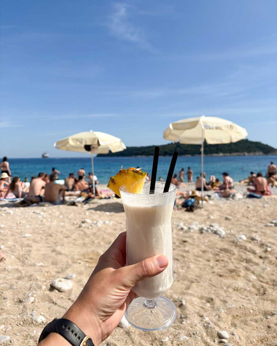 Jesse holds a Pina Colada up in front of the ocean at Banje Beach in Dubrovnik