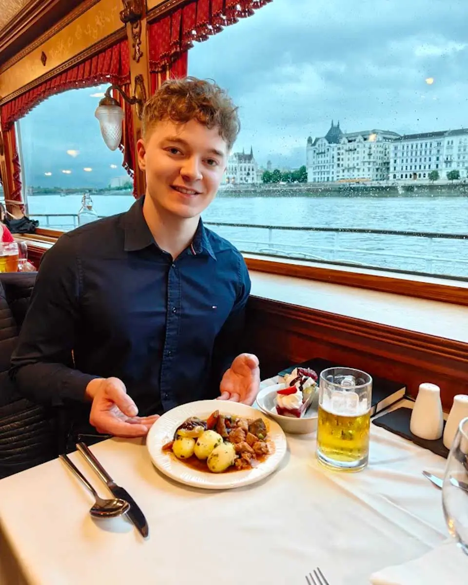 Luke enjoying dinner aboard a Danube River cruise with views of Budapest's waterfront through the window